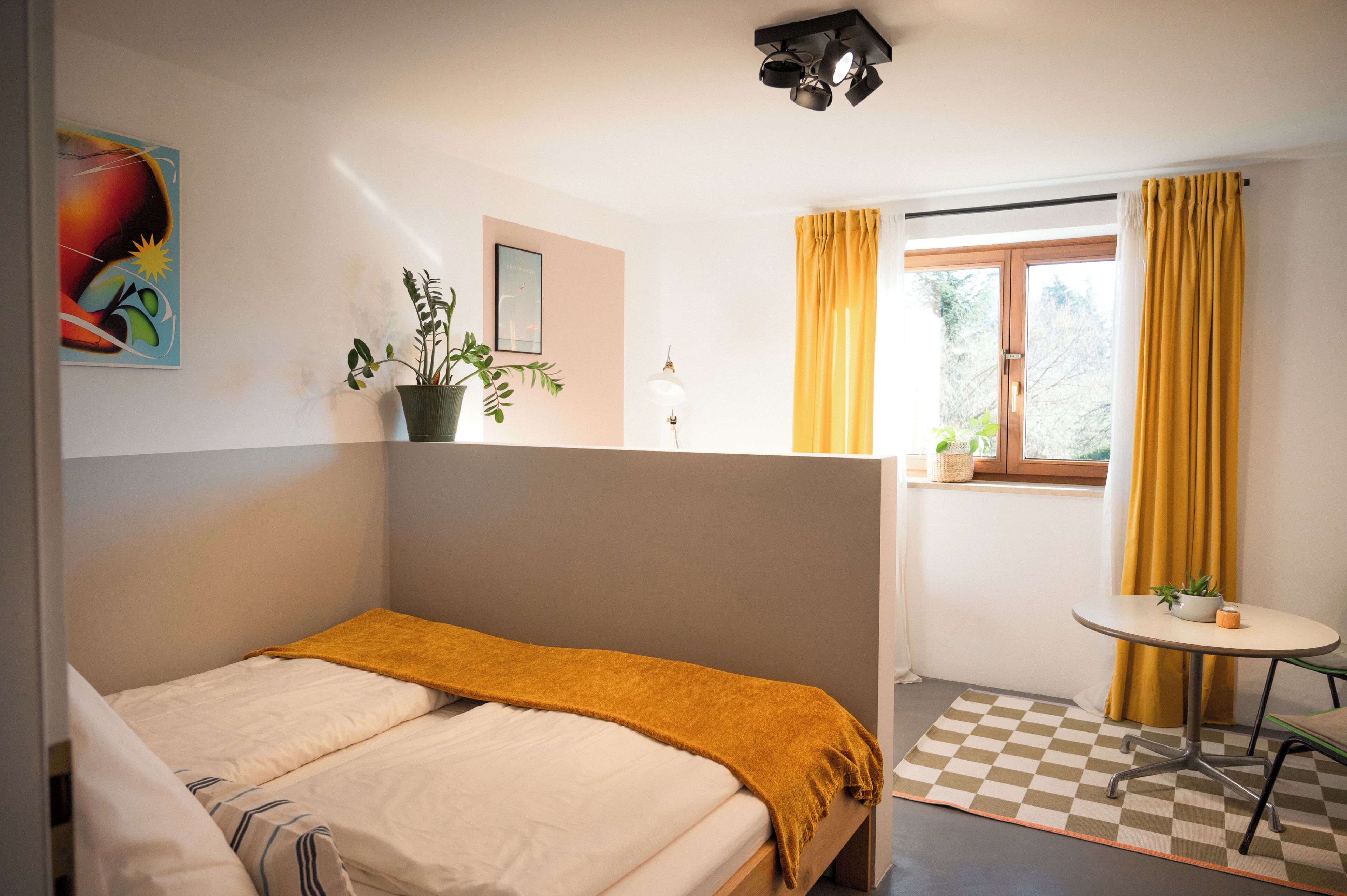 A bedroom with a bed, a small table with a plant, a window with curtains, and a wall with a painting.