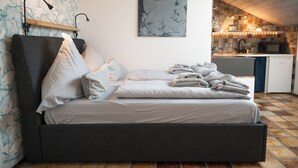 Una cama con almohadas y mantas, una lámpara de noche, una obra de arte colgada en la pared y una zona de cocina con microondas y fregadero.