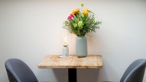 Una pequeña mesa con un jarrón de flores y una vela, dos sillas grises y una pared lisa.