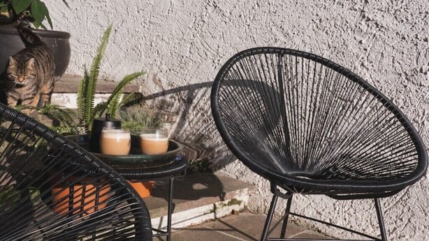 Un patio con dos sillas de alambre negras, una mesa redonda pequeña con dos bebidas, y una planta en maceta.