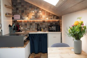 A compact kitchen with a modern design, including a built-in oven, a microwave, and a sink. There is a small dining area with a table and a vase of flowers. - Pura's Haus am Chiemsee: Klein & Einfach (Breitbrunn am Chiemsee)