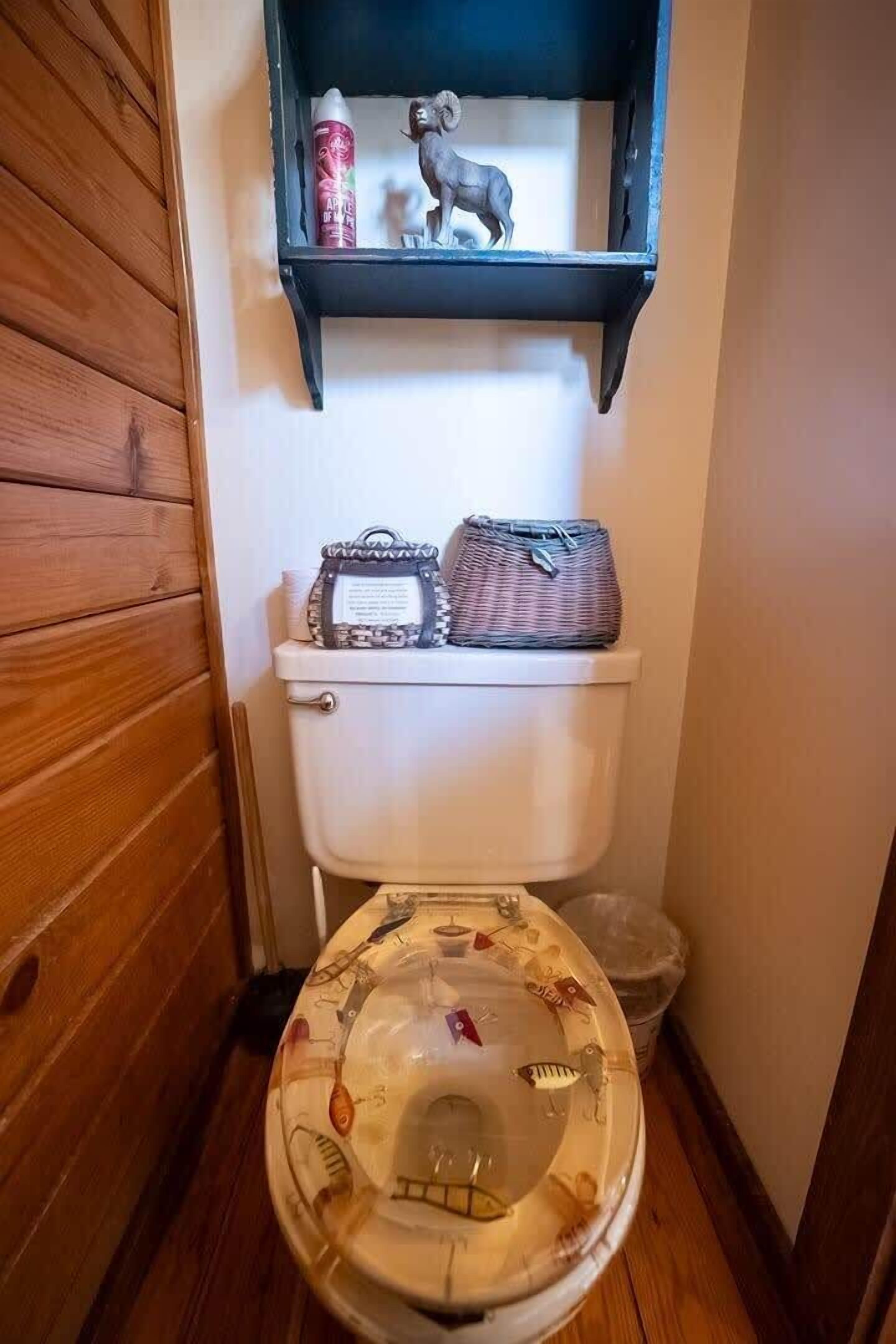 A bathroom with a wooden floor, a white toilet, and a shelf above the toilet holding a small figurine and a can.