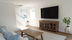 Una sala de estar con un sofá blanco, una mesa de centro de madera, un televisor montado en la pared, un mueble de madera y una planta en maceta.