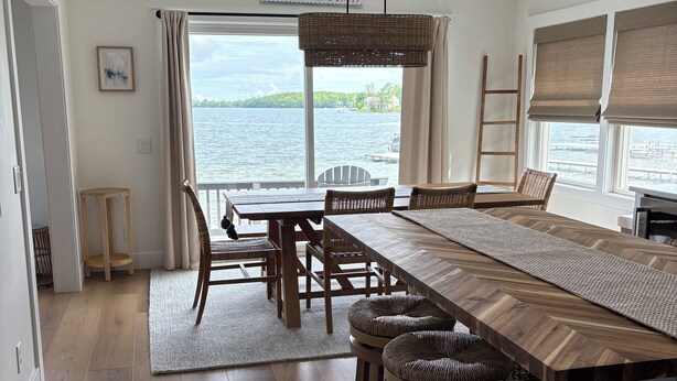 Un comedor con una larga mesa de madera y taburetes de mimbre, un lámpara colgante y vistas al lago.