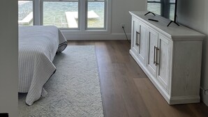 Un dormitorio con un ventanal grande que da a una extensión de agua, una cama con colcha blanca, un armario blanco con televisor y un ventilador de techo.