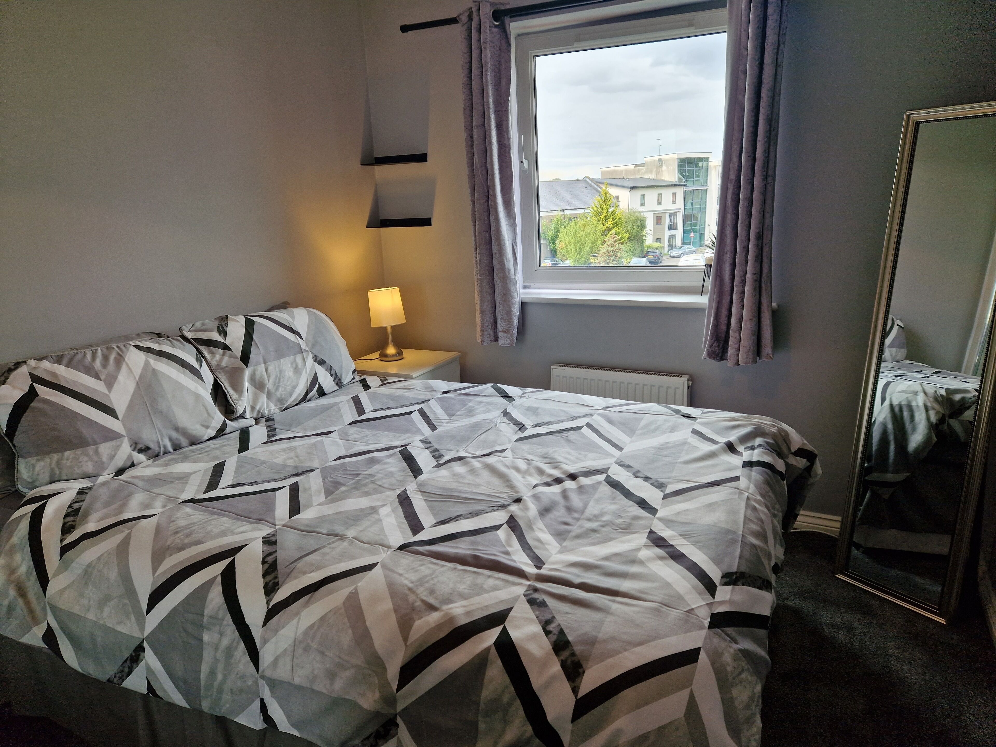 A neatly made bed with a geometric patterned bedspread, a bedside lamp, a window with curtains, and a mirror on the wall.