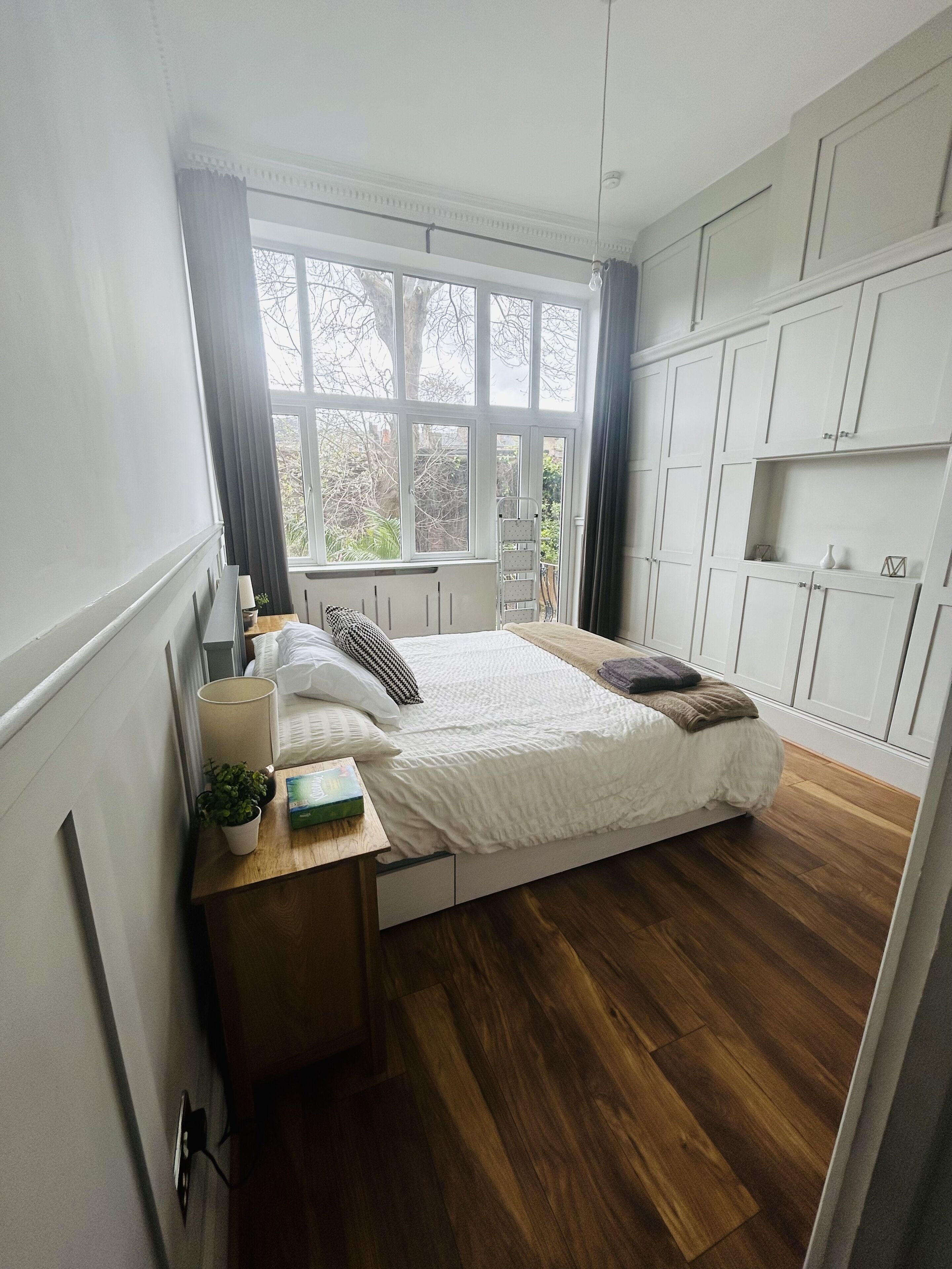 Ein Schlafzimmer mit einem Bett, einem Nachttisch mit Lampe, einer Zimmerpflanze und einem Fenster mit Vorhängen.