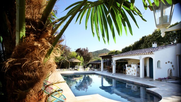 A swimming pool with a curved edge, surrounded by a patio area with outdoor furniture and a white building with stone accents. - Magnifique Villa avec Piscine/Jacuzzi tout confort (Nans-les-Pins)