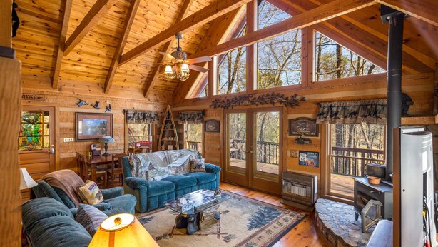 A cozy cabin interior with a wooden ceiling, large windows, a fireplace, and a comfortable seating area.