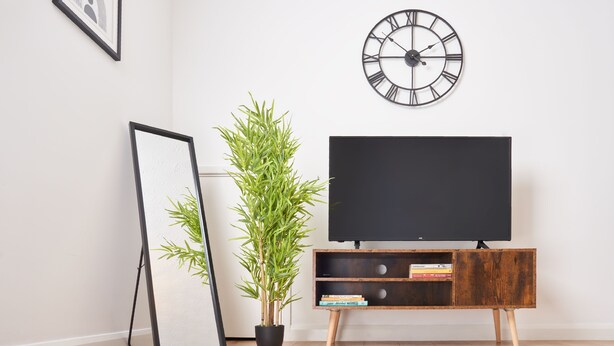 Ein Wohnzimmer mit einem Flachbildfernseher auf einem Holzständer, einer Bambuspflanze im schwarzen Topf, einem gerahmten Bild, das an der Wand lehnt, und einer großen Wanduhr.