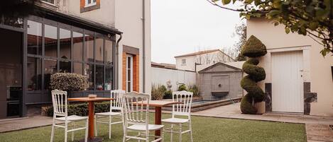 A modern outdoor patio with a wooden table and white chairs, surrounded by a well-maintained lawn and a tall, neatly trimmed hedge.
