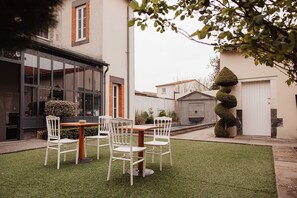 Une terrasse extérieure moderne, avec une table en bois et des chaises blanches, entourée d’une pelouse bien entretenue et d’une haie haute et soigneusement taillée.