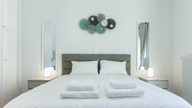 Un lit bien fait, avec du linge de lit blanc et deux serviettes pliées, une tête de lit grise, des tables de chevet avec des lampes et un élément décoratif mural au-dessus du lit.