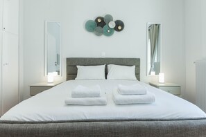 A neatly made bed with white linens and two folded towels, a grey headboard, bedside tables with lamps, and a wall decoration above the bed. - Shades of Athens (Athens)