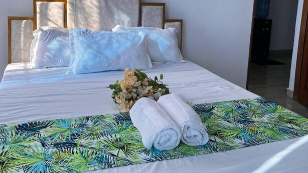 Ein ordentlich bezogenes Bett mit weißen Laken, einer Bettdecke mit tropischem Blattmuster und einem Blumenstrauß.