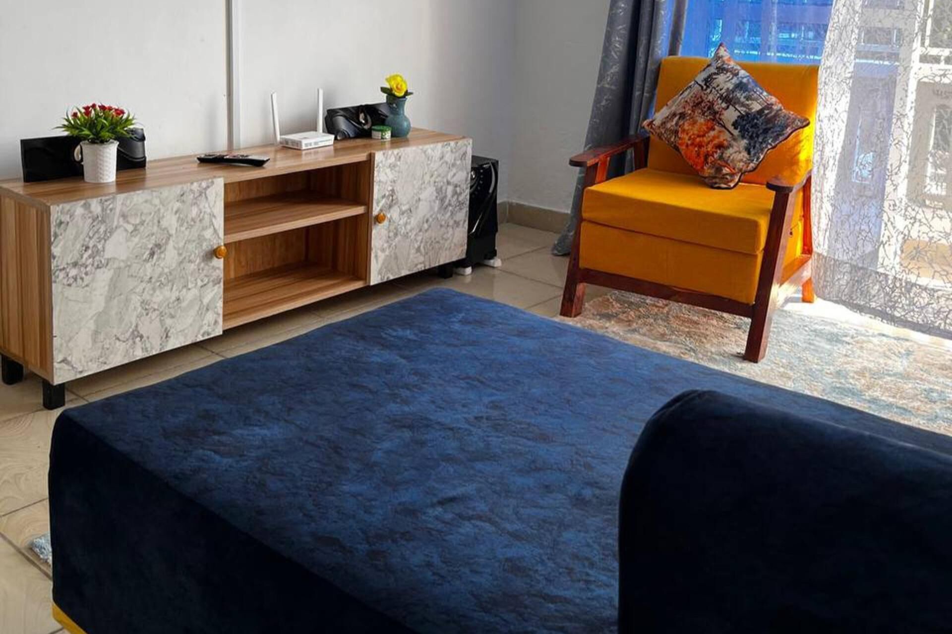 A living room with a blue sofa, a wooden cabinet, a yellow armchair with a colorful cushion, a small table with a vase and a plant, and a window with a view.