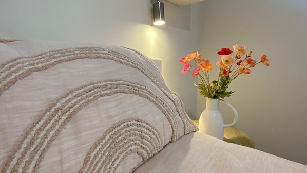 A bed with a beige blanket and a white vase with flowers on a bedside table.