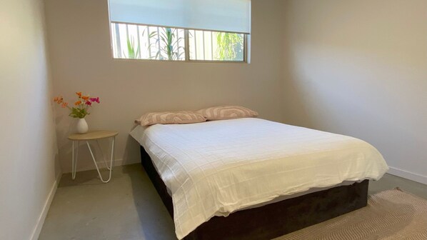 A bedroom with a bed, a small table with a vase of flowers, and a window with a view of greenery. - Charming Beach Getaway -Tucked away in Valla beach is the charming home that has a unique private ba (Valla Beach)