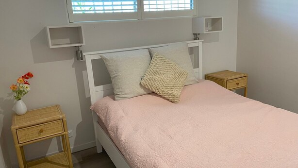 Ein Schlafzimmer mit einem Bett, zwei Nachttischen, einem Fenster mit Jalousien und einer Vase mit Blumen.