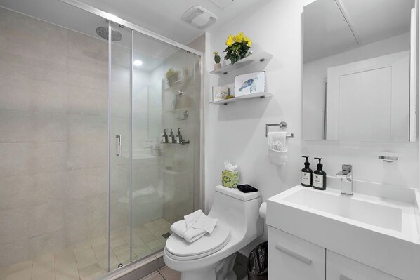 Un baño moderno con ducha envuelta en vidrio, inodoro blanco y lavamanos con artículos de aseo.