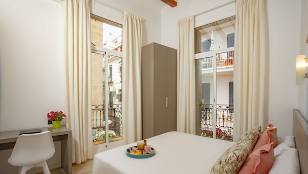 A bedroom with a bed, a desk, and a window with curtains. - Antonios Hostal (Sitges)