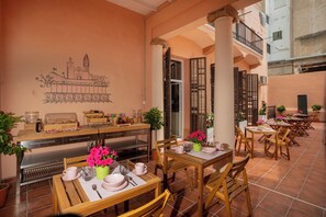 A cozy outdoor dining area with wooden tables and chairs, a mural of a building on the wall, and potted plants.