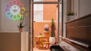 A piano, a neon flower sign, a dining table with chairs, and potted plants. - Antonios Hostal (Sitges)