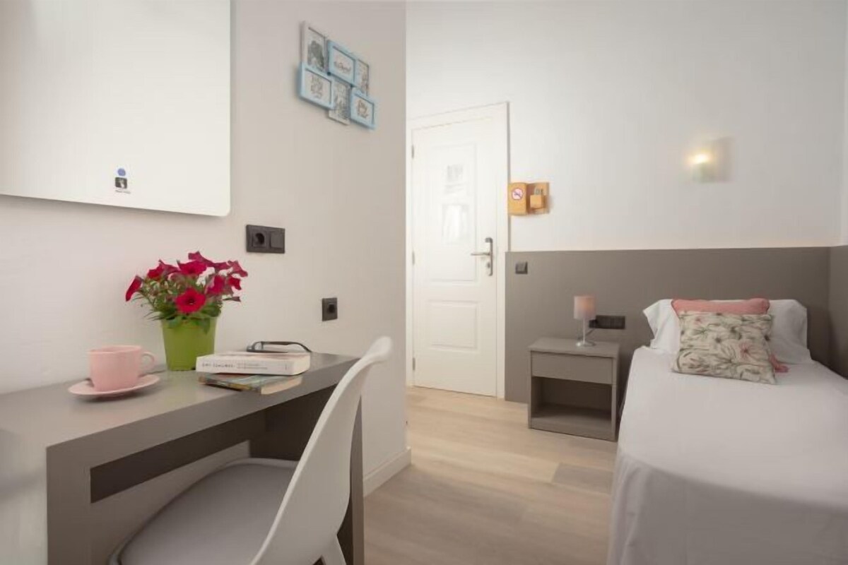 Et moderne hotelværelse med et skrivebord, stol og seng med sengegavl. Der er et lille natbord med lampe og en vægmonteret hylde.