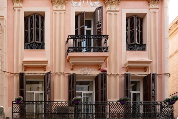 A pink building with three balconies, each with a wrought iron railing and potted plants.
