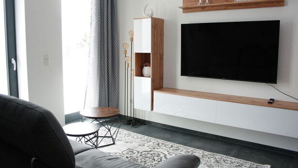 Un salon moderne avec un téléviseur à écran plat fixé au mur, une étagère en bois au-dessus et une petite table ronde avec une chaise.