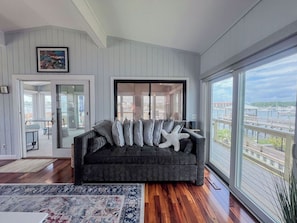A modern living room with a dark grey sofa, a large window with a city view, and a framed picture on the wall.
