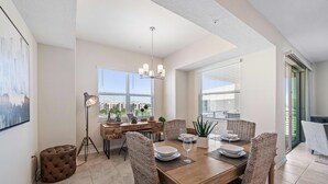 A dining area with a wooden table set for four, surrounded by wicker chairs. A floor lamp and a tufted ottoman are visible. - Pool View 2 bedroom! Close to Disney World! (Kissimmee)