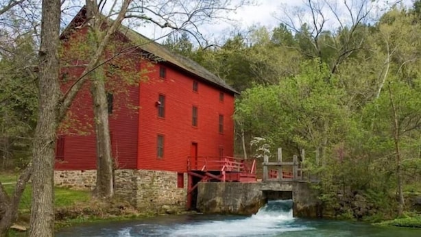 Un moulin rouge avec un petit pont enjambant une rivière, le tout entouré d’arbres.