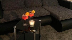 A modern living room with a dark grey sectional sofa, a small black side table with two red cocktails and a lit candle, and a white wall.