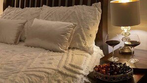 A bed with white linens and pillows, a bedside table with a lamp and two wine glasses, and a bowl of fruit.