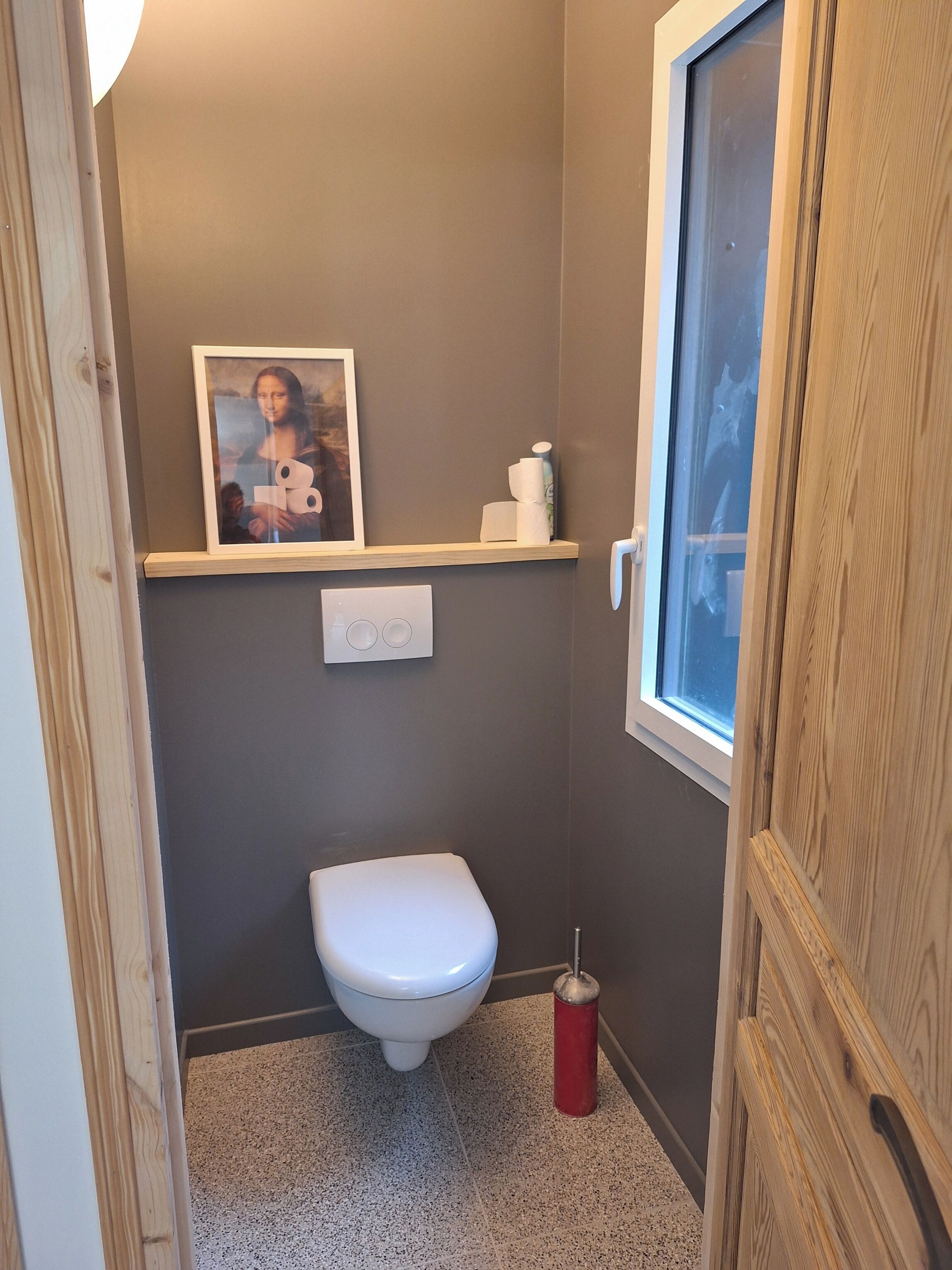 A compact bathroom with a toilet, a small shelf with toiletries, a framed picture on the wall, and a window.
