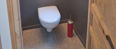 A compact bathroom with a toilet, a small shelf with toiletries, a framed picture on the wall, and a window.
