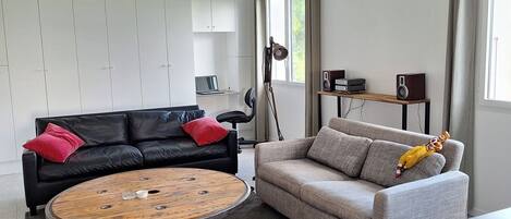 Una sala moderna con un sofá de cuero negro, un sofá gris, una mesa de centro redonda de madera y una pequeña mesa auxiliar de madera con un altavoz.