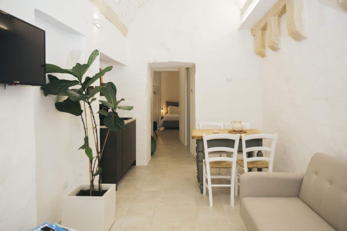 Una sala de estar moderna con un sofá, una mesa de comedor y sillas. Hay un televisor montado en la pared y una planta en maceta.