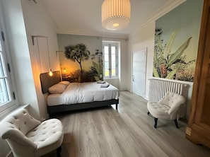 Ein Schlafzimmer mit Bett, zwei Stühlen, einem Heizkörper, einem Fenster und Wandbild.