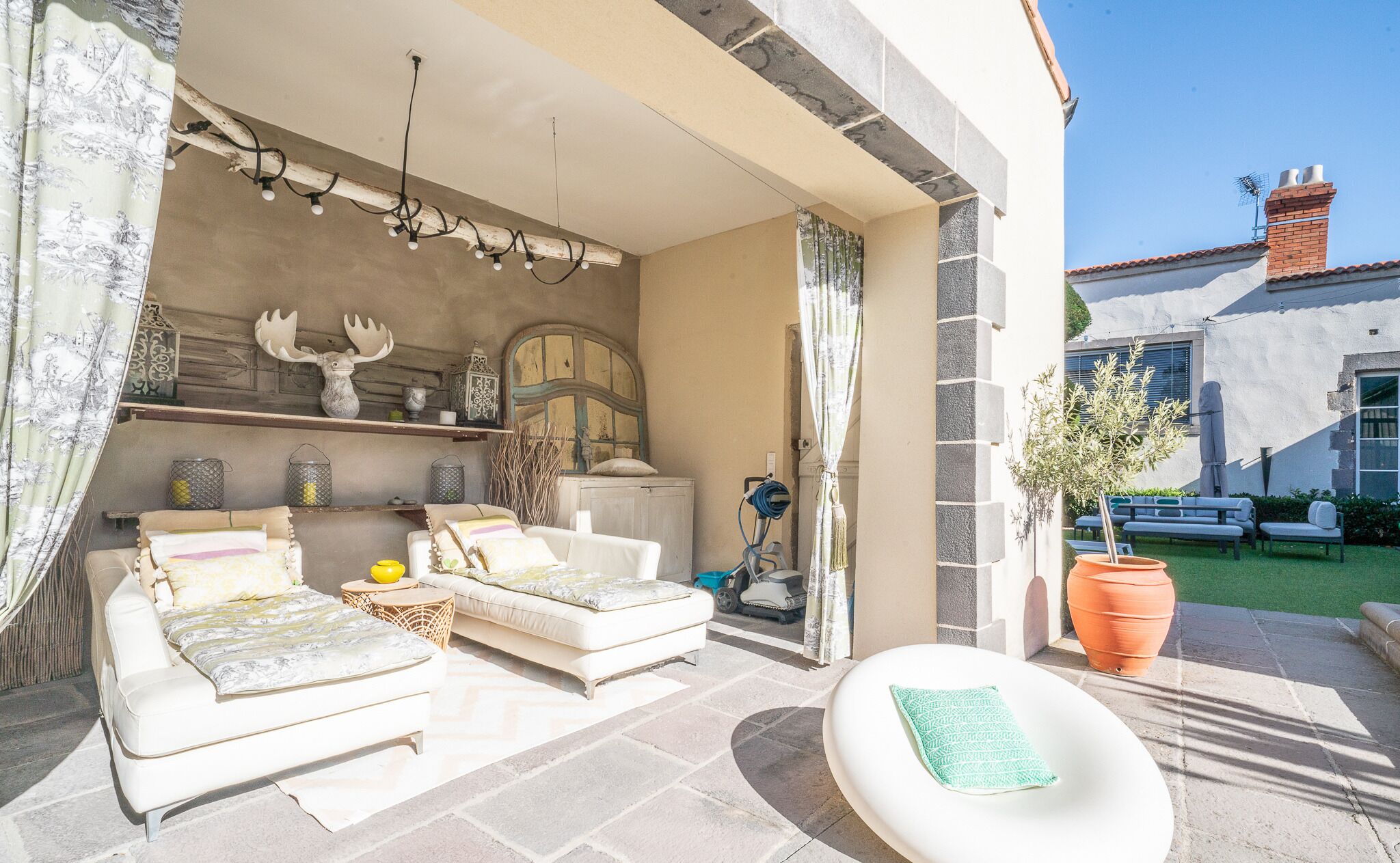 Un espace extérieur accueillant avec un canapé, un fauteuil et une petite table. Un mur décoratif au style rustique et un pot de fleurs agrémentent l’ensemble.