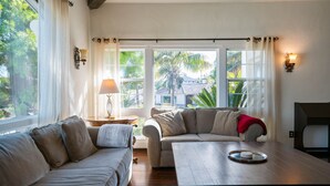 Uma sala de estar aconchegante com dois sofás, uma mesa de centro de madeira e uma luminária. Janelas amplas com cortinas revelam um visual com vegetação e uma casa lá fora.