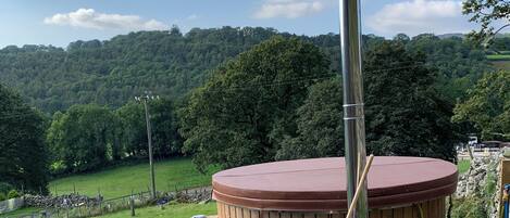A hot tub with a wooden deck and a chimney, surrounded by a gravel area and greenery.