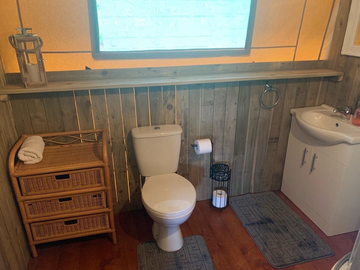 Ein kleines Badezimmer mit Toilette, einem Korbschrank, einem Waschbecken und einem Fenster über der Toilette.