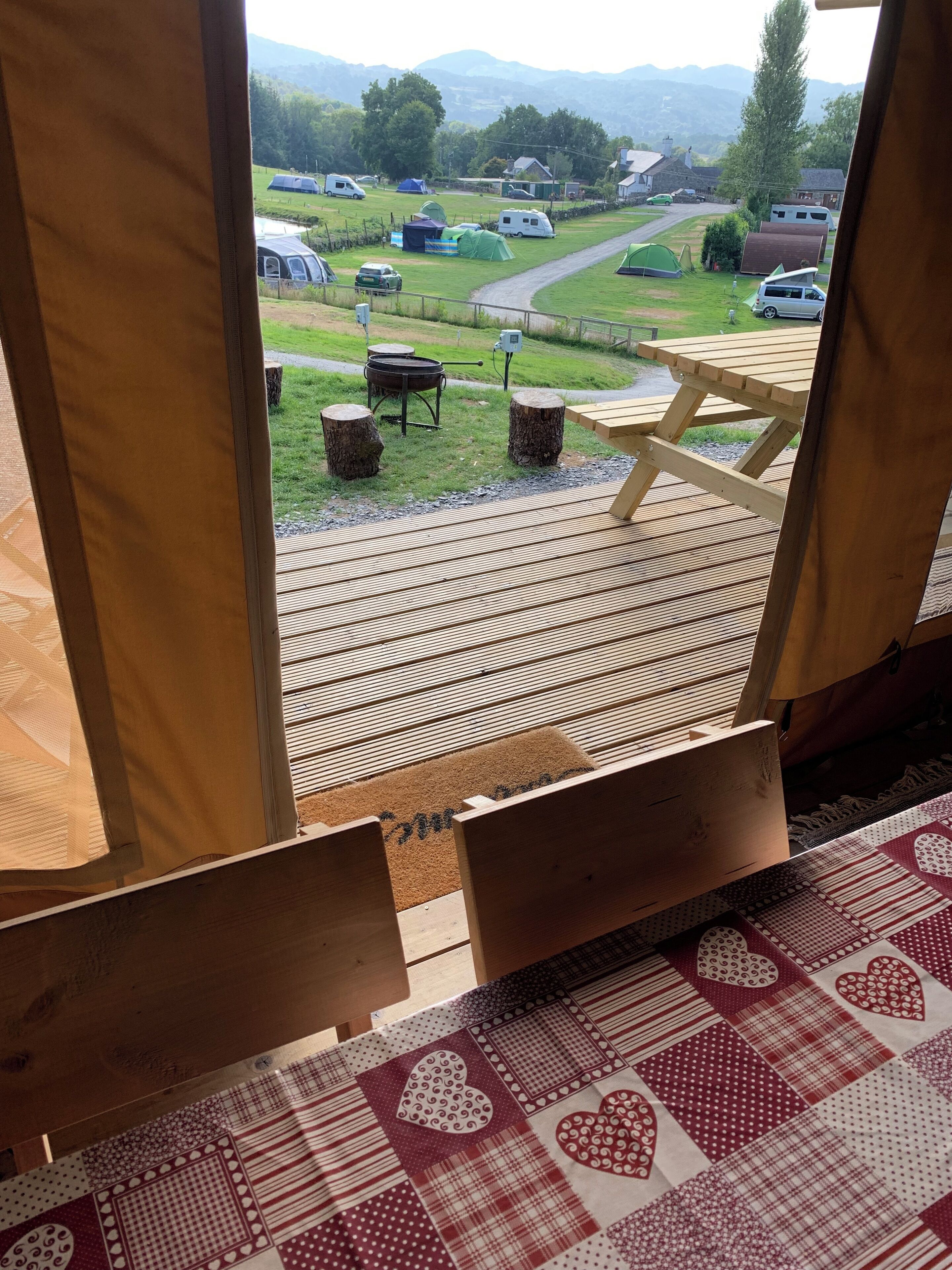 A view from inside a cabin with a wooden deck, a picnic table, and a doormat with a heart pattern.