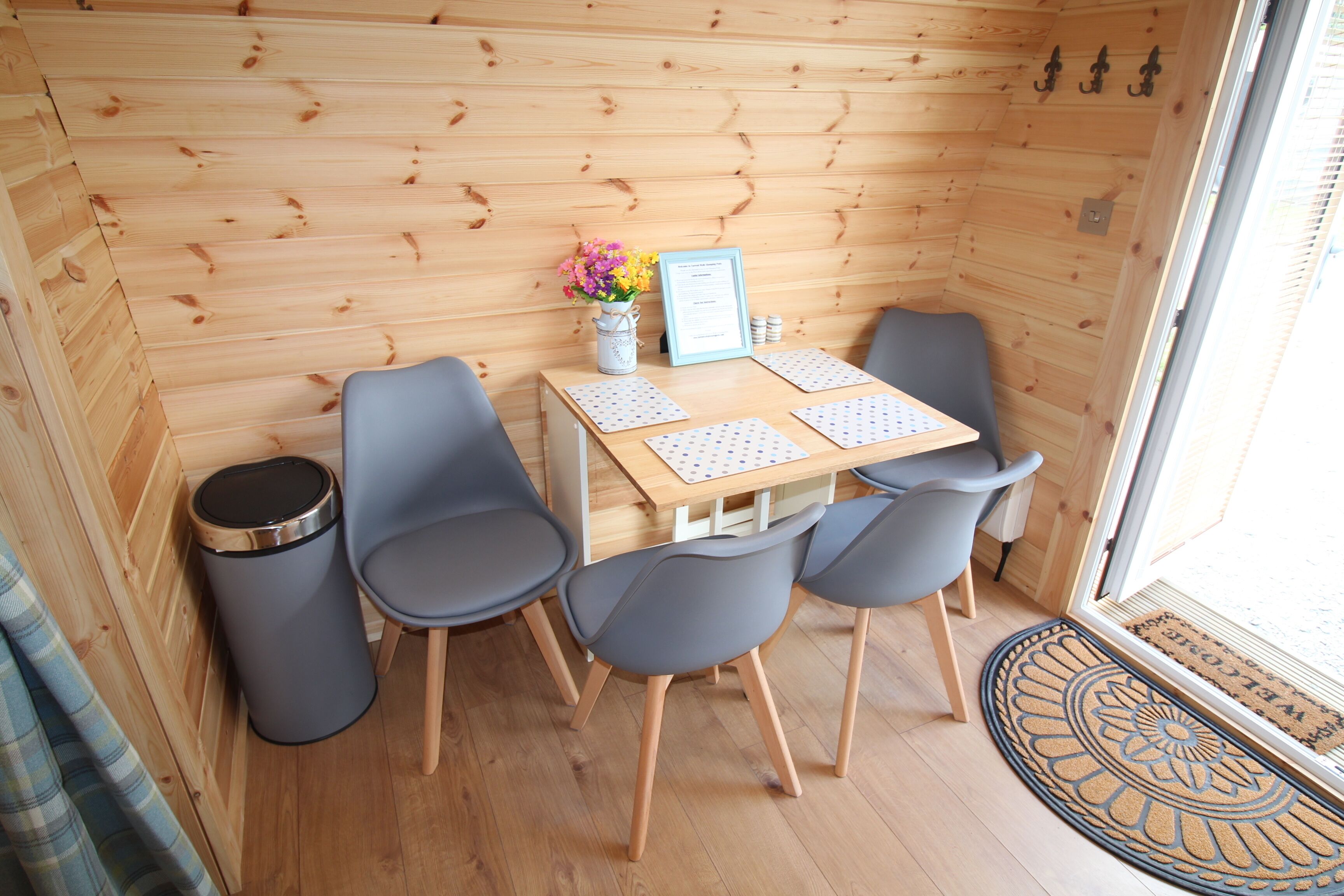 Una habitación pequeña de madera con una mesa de madera, cuatro sillas, un cubo de basura y una puerta con felpudo.