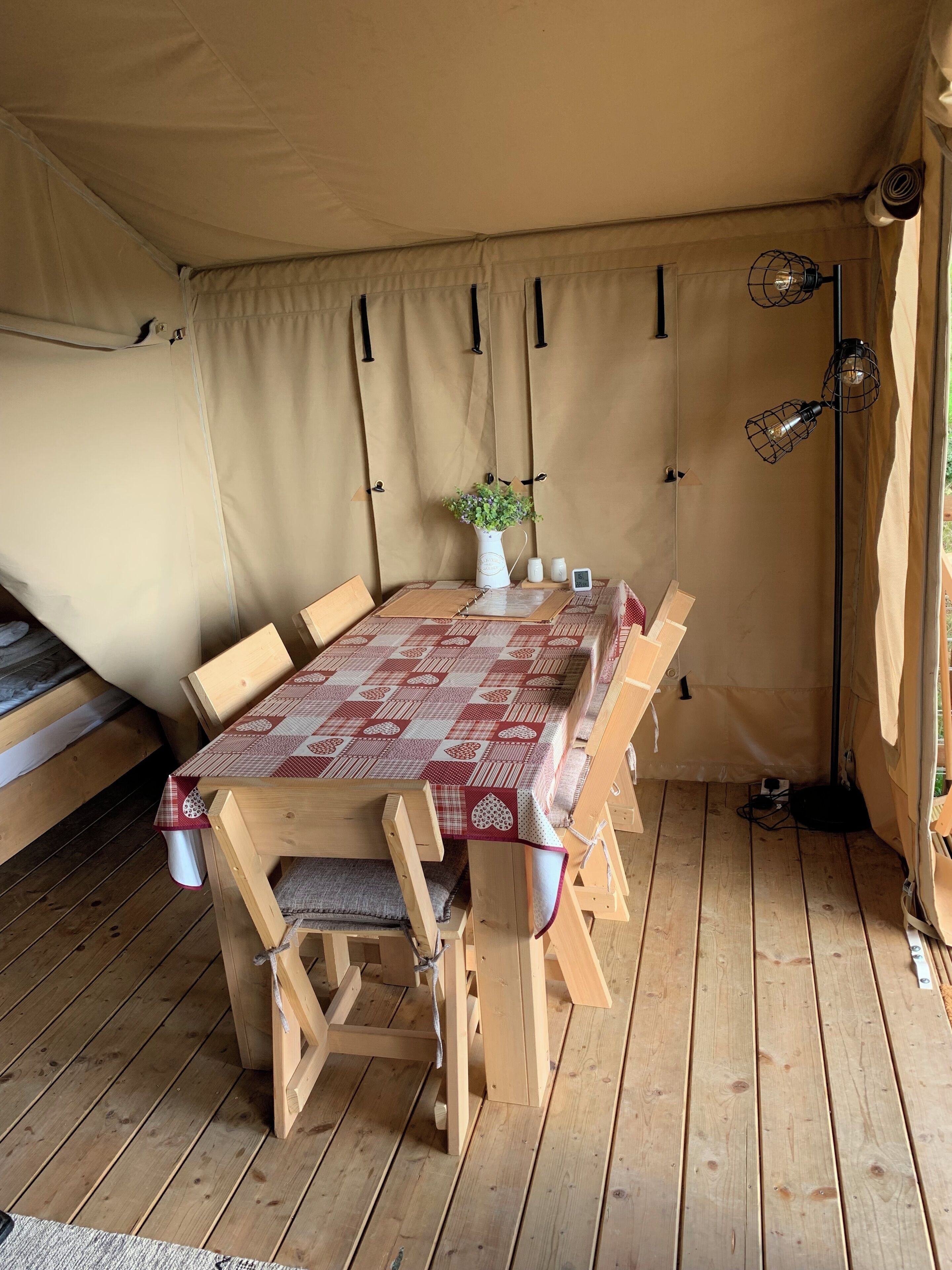 Een houten tafel met geruit tafelkleed, omringd door stoelen, onder een tent met beige wanden en een hanglamp.