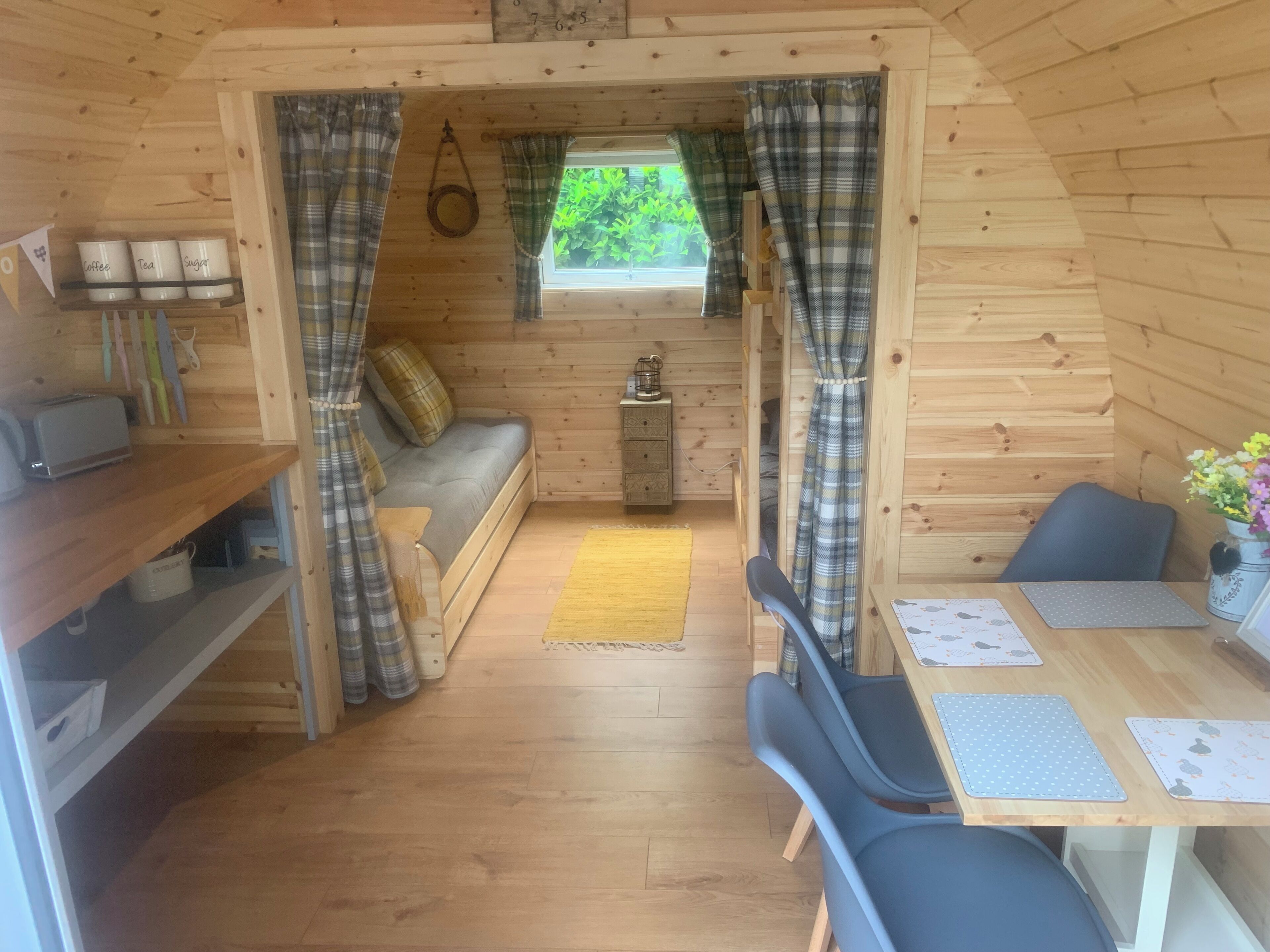 L’intérieur d’un chalet accueillant, avec un décor en bois, un canapé, une table à manger entourée de chaises et une petite fenêtre agrémentée de rideaux.