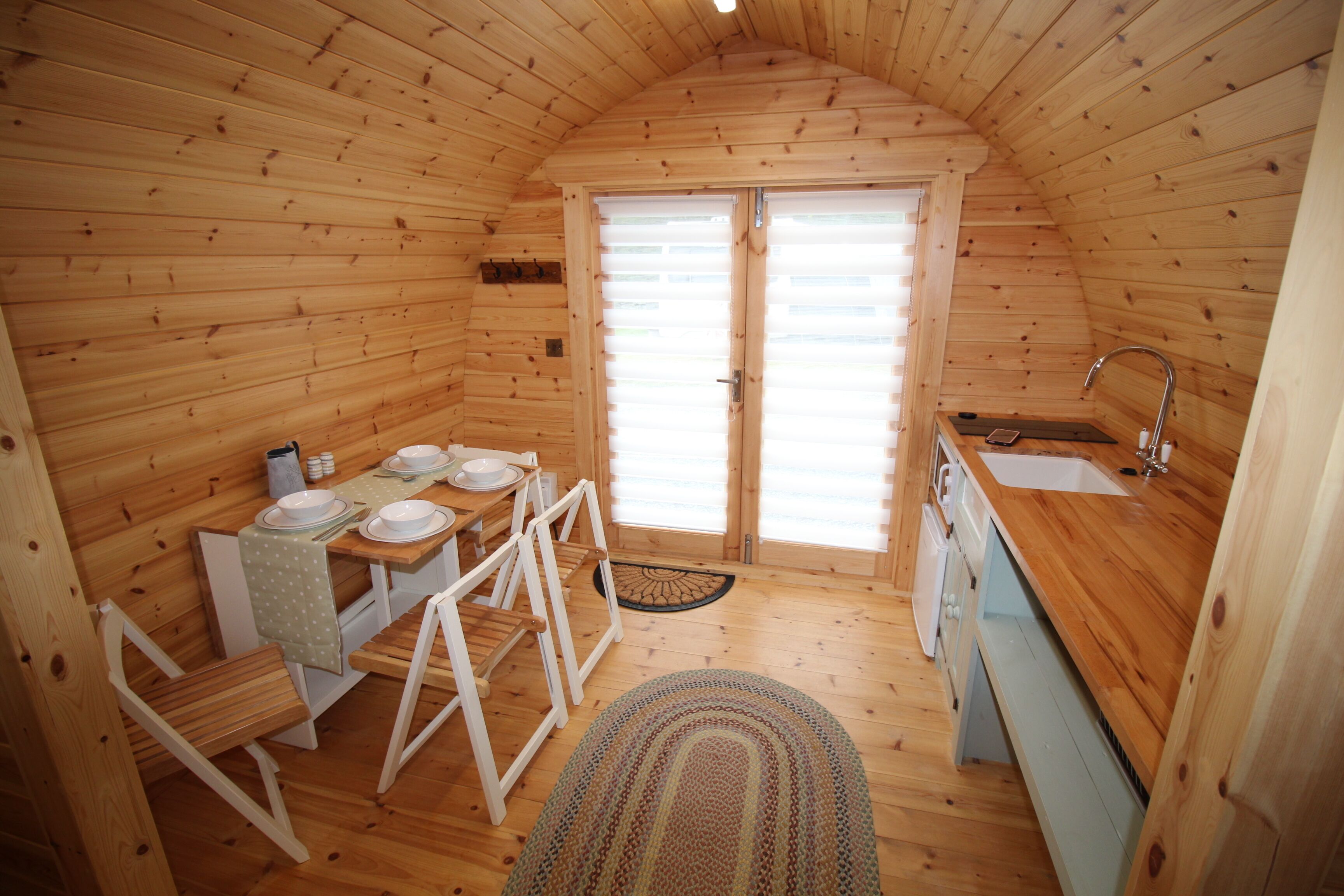 Un piccolo chalet in legno con zona pranzo, cucina dotata di lavello e porta che dà all'esterno.