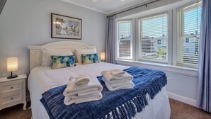 A neatly made bed with folded towels, a ceiling fan, a framed picture on the wall, and a window with blinds.